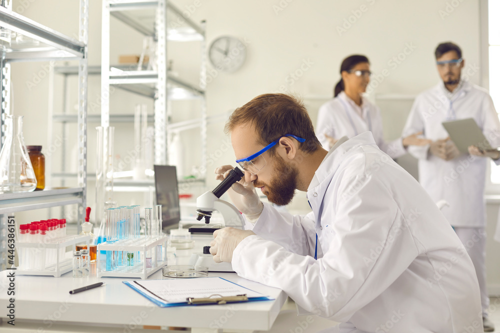 Professional male scientist looking through microscope in scientific research laboratory. Genetic engineer workplace. Colleague team on background. Technology, medicine and science concept. Professional male scientist looking through microscope in scientific research laboratory. Genetic engineer workplace. Colleague team on background. Technology, medicine and science concept.