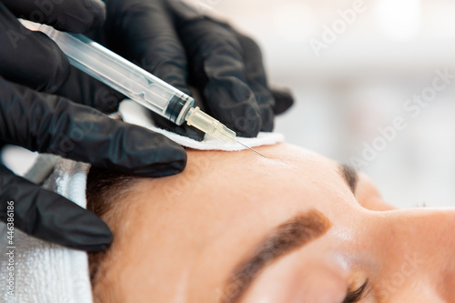 Close-up of female s head getting injection at her forehead in the cosmetology salon. Cosmetologist in latex gloves with syringe injects a medicine. Concept of plastic surgery and face lifting