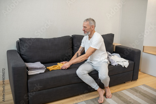 A gray-haired man sitting on the sofa and looking at the clothes lying near