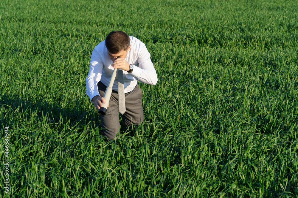 businessman poses with a spyglass, he looks into the grass and looks for something, green grass and blue sky as background businessman poses with a spyglass, he looks into the grass and looks for something, green grass and blue sky as background