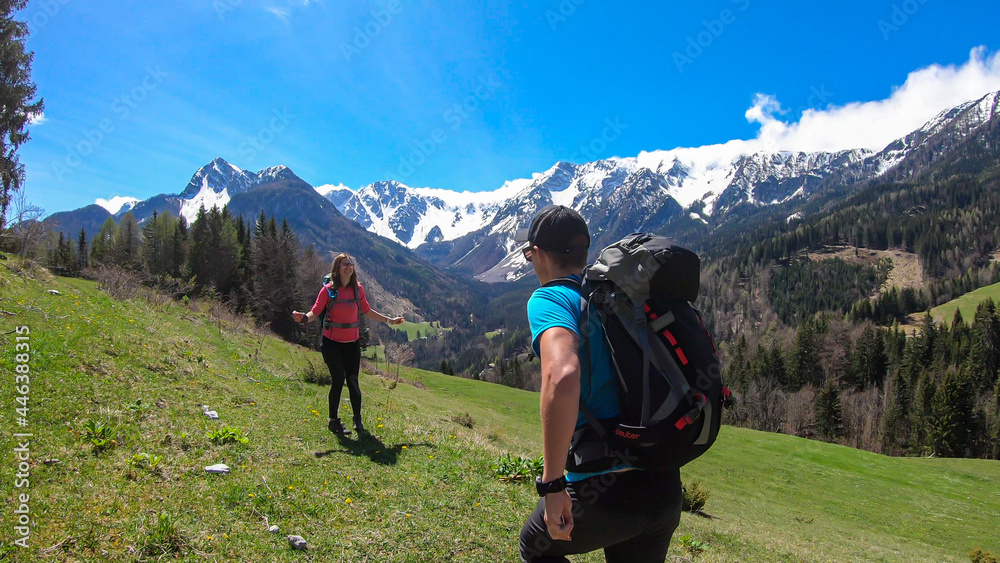 A couple with a big hiking backpacks dancing with the panoramic view on Baeren Valley in Austrian Alps. The highest peaks are snow-capped. Lush green pasture. Clear and sunny day. High mountain chains