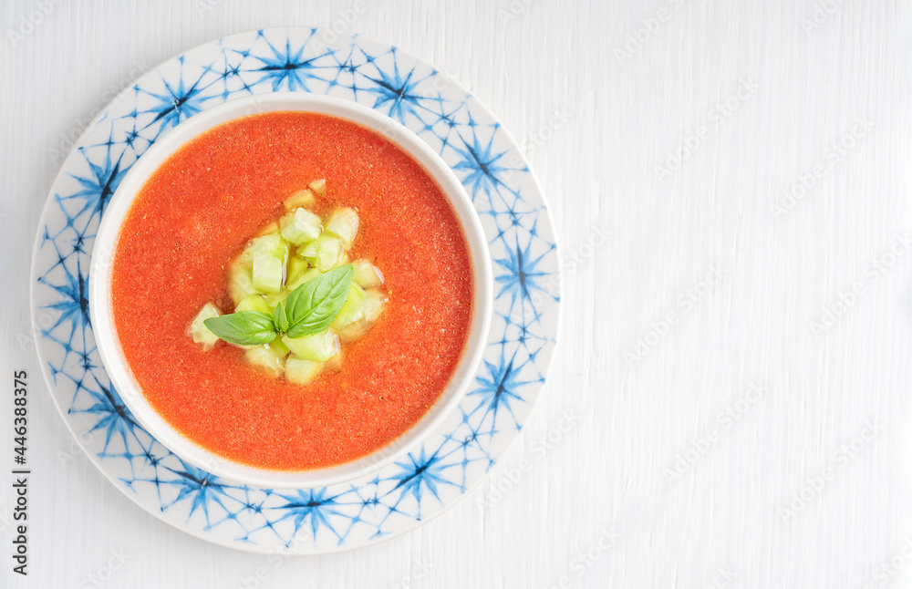 Top view of spanish gazpacho cold refreshing tomato soup made of raw blended vegetables served in bowl on plate with basil leaves and cucumber cubes on white wooden background. Image with copy space Top view of spanish gazpacho cold refreshing tomato soup made of raw blended vegetables served in bowl on plate with basil leaves and cucumber cubes on white wooden background. Image with copy space