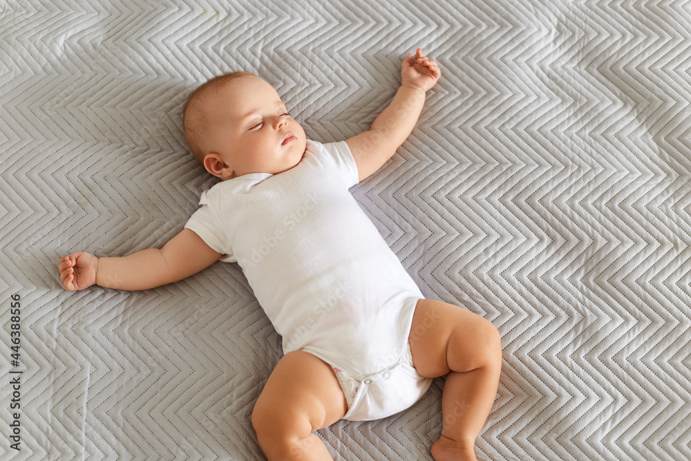 Naklejka premium Charming cute baby lying on a bed, sleeping in a bright room on grey blanket, laying with spreading hands, afternoon sleep indoor.