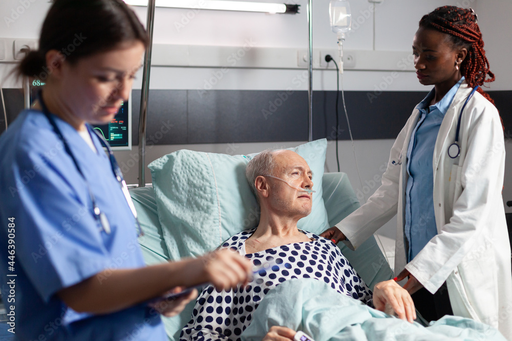 African doctor ward in hospital room discussing with sick senior man laying in bed breathing through oxygen tube with iv dirp bag attached, during consultation. Medical nurse taking notes on clipboard African doctor ward in hospital room discussing with sick senior man laying in bed breathing through oxygen tube with iv dirp bag attached, during consultation. Medical nurse taking notes on clipboard