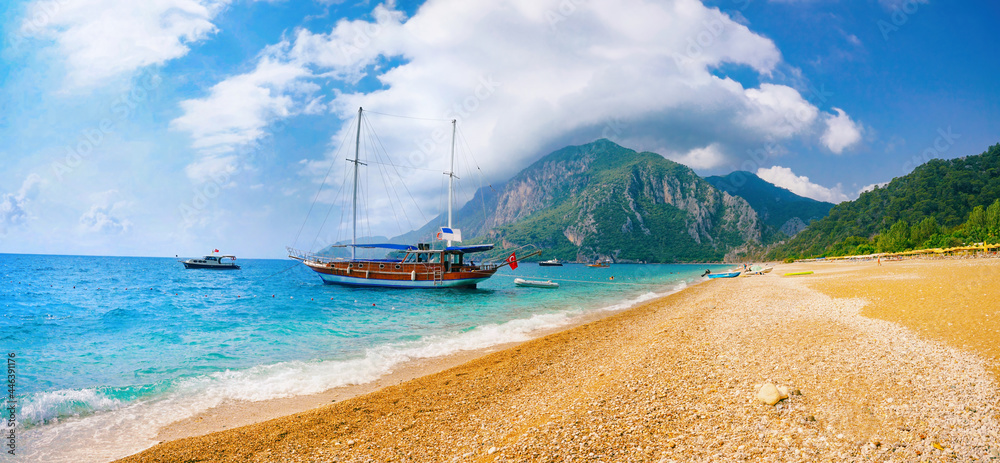 Fototapeta premium Beautiful panoramic view of sea and beach Cirali, Kemer, Antalya, Turkey. Ship against backdrop of mountains and blue sky with clouds on sunny day.