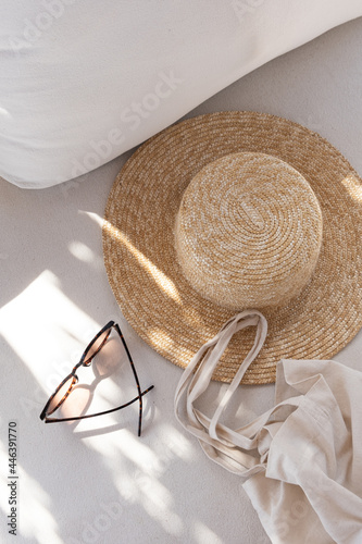 Flatlay of women's fashion accessories. Stylish female sunglasses, straw hat, shopper bag on white lounge couch with pillows. Top view