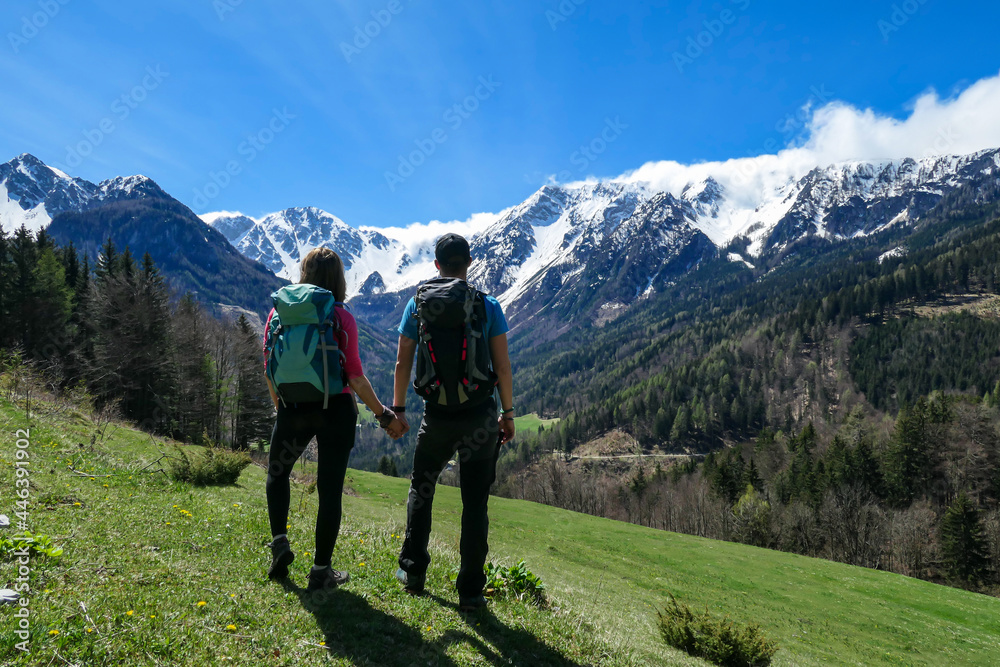 A couple with big hiking backpacks holding hands and enjoying the panoramic view on Baeren Valley in Austrian Alps. The highest peaks are snow-capped. Lush green pasture. Sunny day. High mountains
