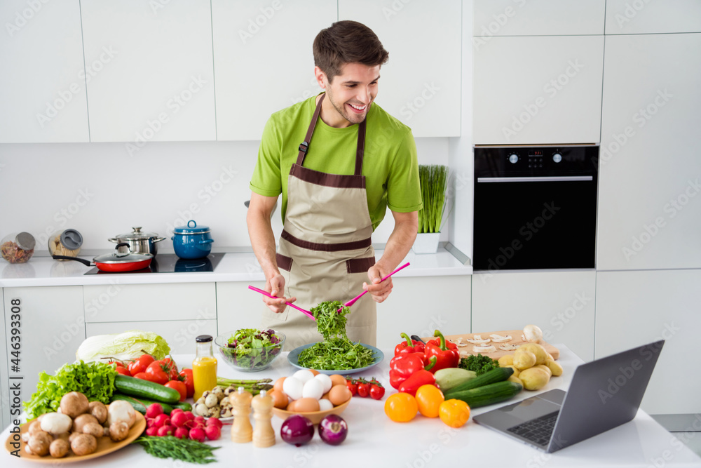Photo portrait young man in apron cooking vegetables salad with laptop watching recipe video on youtube