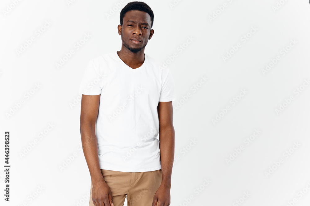 a young man of African appearance in beige trousers and a white T-shirt looks forward on a light background a young man of African appearance in beige trousers and a white T-shirt looks forward on a light background