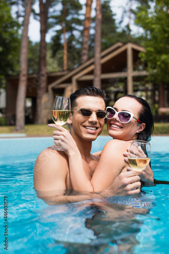 Young couple in sunglasses smiling at camera while holding white wine in pool