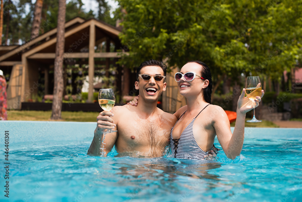 Cheerful couple in sunglasses holding glasses of wine in swimming pool
