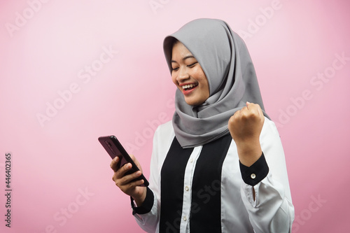 Beautiful young asian muslim woman smiling confident, enthusiastic and cheerful with hands holding smartphone, hands clenching, punching, victory, success, good work, isolated on pink background © MunirSr