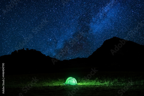 Tent glowing under a starry summer night in the countryside. Tent illuminated by a light. Camping in the mountains under the starry sky. Camping and wild life concept. Real outdoor adventure.