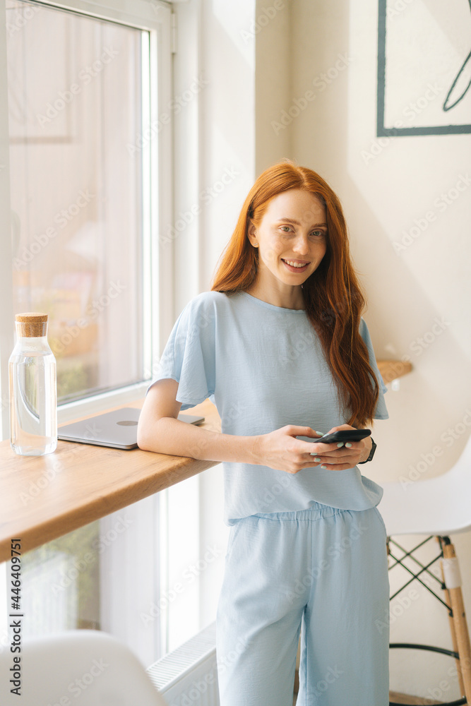 Medium shot vertical portrait of smiling attractive young woman using phone , typing online messages standing by window in cafe. Pretty redhead Caucasian lady having leisure activity in coffee shop. Medium shot vertical portrait of smiling attractive young woman using phone , typing online messages standing by window in cafe. Pretty redhead Caucasian lady having leisure activity in coffee shop.