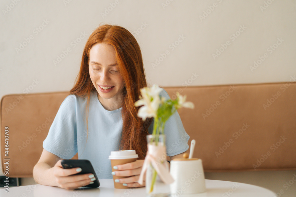 Front view of smiling young woman using smartphone chatting with friends and drinking hot coffee sitting at desk in cozy cafe. Pretty redhead Caucasian lady having leisure activity in coffee shop. Front view of smiling young woman using smartphone chatting with friends and drinking hot coffee sitting at desk in cozy cafe. Pretty redhead Caucasian lady having leisure activity in coffee shop.