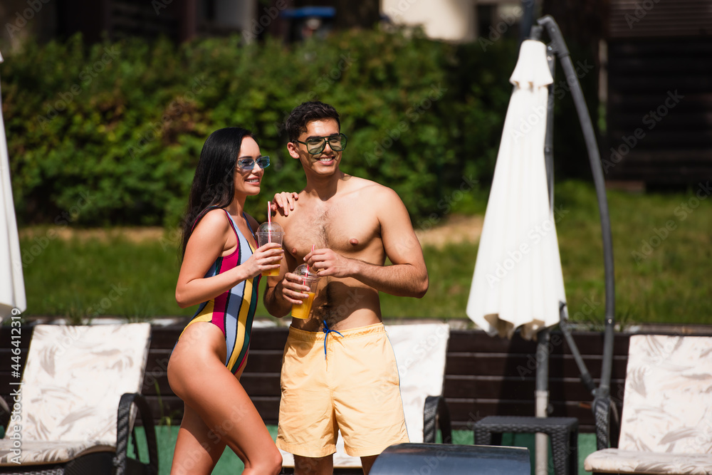 Smiling woman with orange juice touching boyfriend in swim trunks on resort