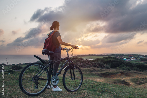 Young woman watching the sunset and the beautiful view of nature while standing with her bicycle on a hill.