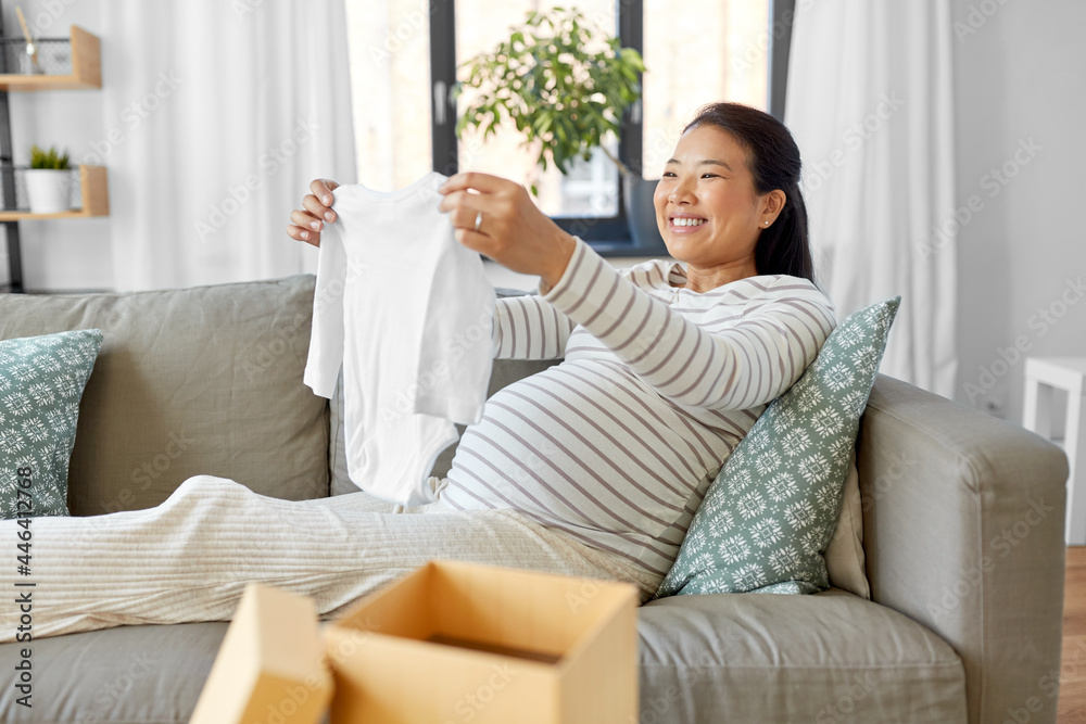 pregnancy, holidays and people concept - happy smiling pregnant asian woman opening gift box with baby's bodysuit at home