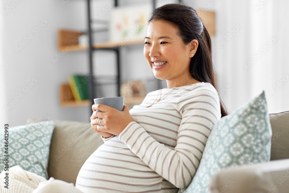pregnancy, rest, people and expectation concept - happy smiling pregnant asian woman with mug sitting on sofa at home and drinking tea