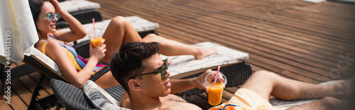 Man in sunglasses holding orange juice near blurred girlfriend on deck chair, banner