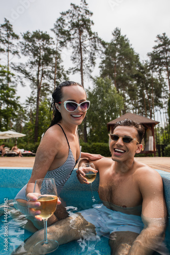 Cheerful woman with blurred wine relaxing near wet boyfriend in swimming pool