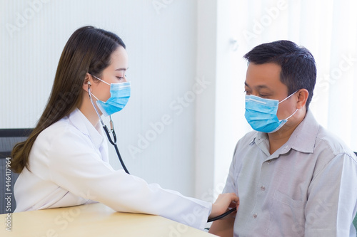 Asian male patient are checked up his health while a woman doctor use a stethoscope to hear heart rate and the sound breathing of hims in Coronavirus pandemic By wearing a surgical mask at all times. 