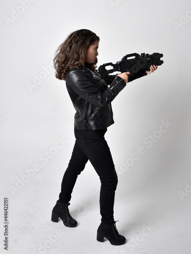 Full length portrait of young woman with natural brown hair, wearing black leather scifi outfit with corset, standing pose holding a gun on light grey studio background.