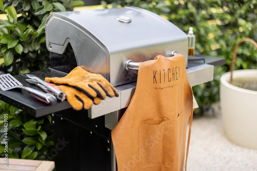 Fotografia Modern gas grill with leather apron and gloves stands at backyard