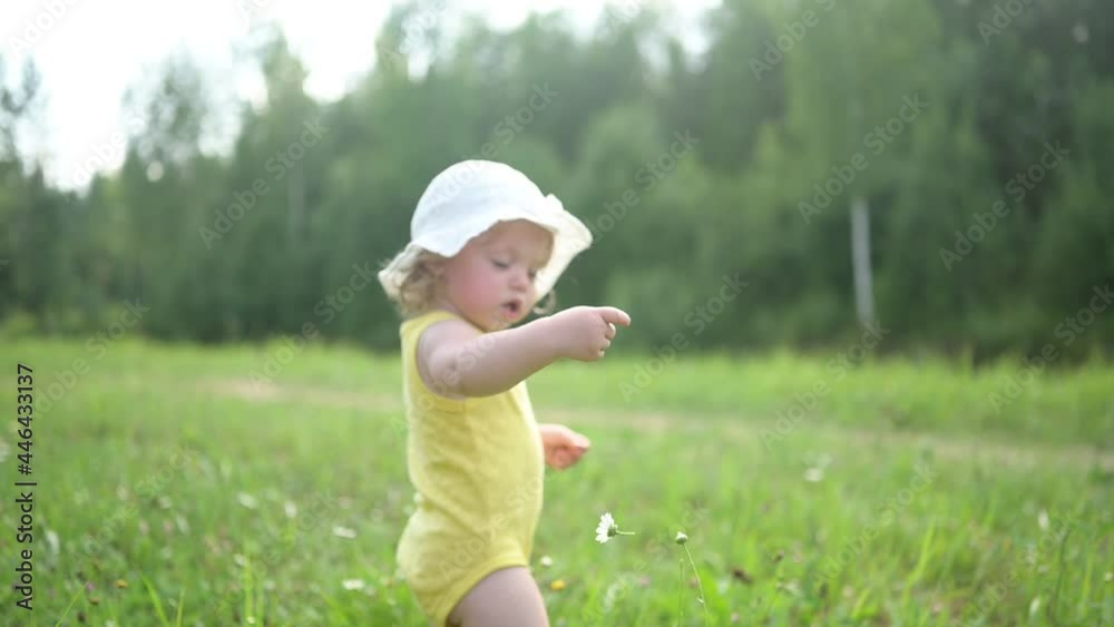 Little funny cute blonde girl child toddler in yellow bodysuit and white hat walking in field with green grass and daisies outside at summer. Baby picks wild flowers. Healthy happy childhood concept Little funny cute blonde girl child toddler in yellow bodysuit and white hat walking in field with green grass and daisies outside at summer. Baby picks wild flowers. Healthy happy childhood concept