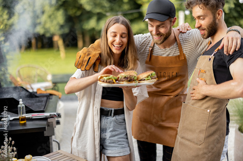 Young people enjoy yummy burgers made on a grill at picnic, standing together and having fun. Friends cooking at backyard outdoors. American lifestyle