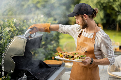 Handsome man in cap and apron making burgers on a grill at backyard. Cooking outdoors and american lifestyle concept