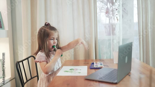 Beatiful girl study online. Girl drawing, painting and distance learning. Baby are sitting at the table and looking at the computer, laptop. Stay home and have fum. Self-isolation. Epidemic.