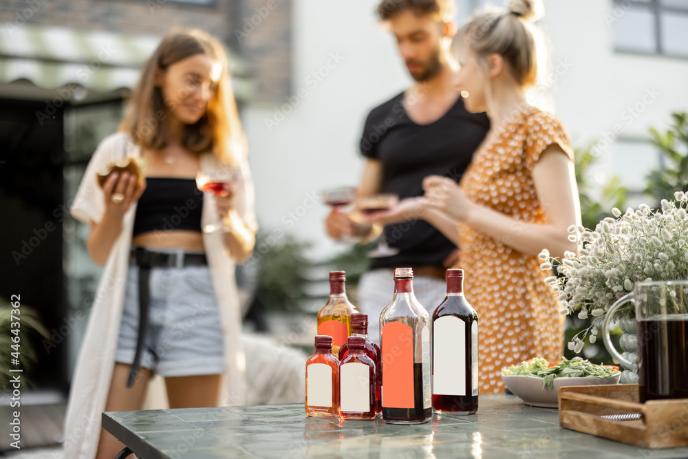 Young people celebrate and have fun, drinking alcohol drinks at backyard. Bottles with blank labels on the table on the foreground Young people celebrate and have fun, drinking alcohol drinks at backyard. Bottles with blank labels on the table on the foreground