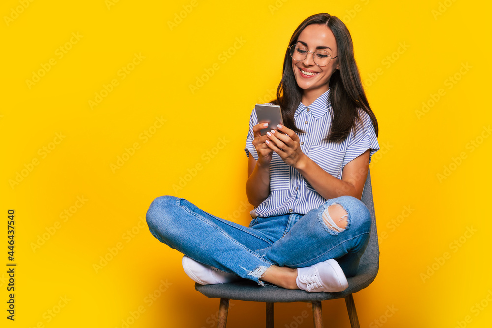 Happy beautiful stylish woman is sitting on the chair and using her smart phone while she is isolated on yellow background Happy beautiful stylish woman is sitting on the chair and using her smart phone while she is isolated on yellow background