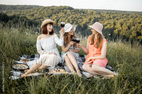 Group of three young stunning women at a picnic in the field. Smiling best friends drinking red wine on the lawn. Girls in stylish dresses and hats relaxing and have a lunch outside. Provence picnic