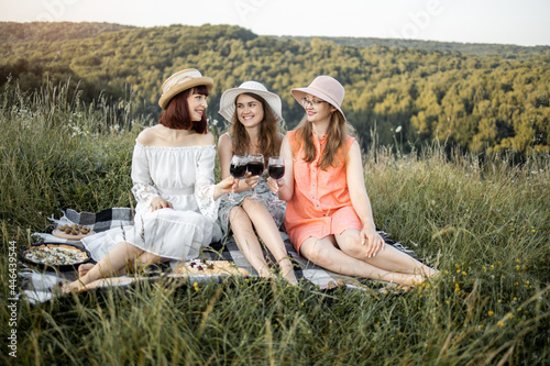 Cheerful female friends have a great time together on a picnic in a picturesque meadow with green hills. Girls in hats and dresses sitting on the blanket and toasting wine glasses