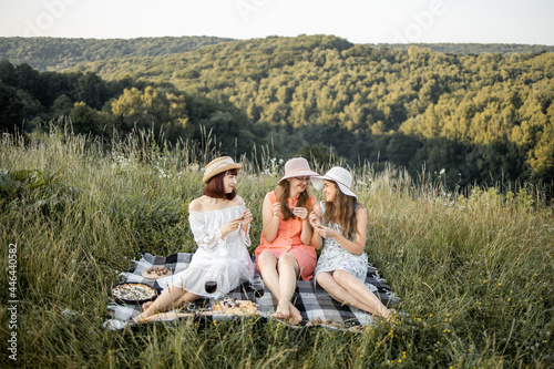 Female friendship. Picnic time. Three attractive girls having fun and enjoying picnic lunch, against the background of green hills. Blanket with food spread on it.