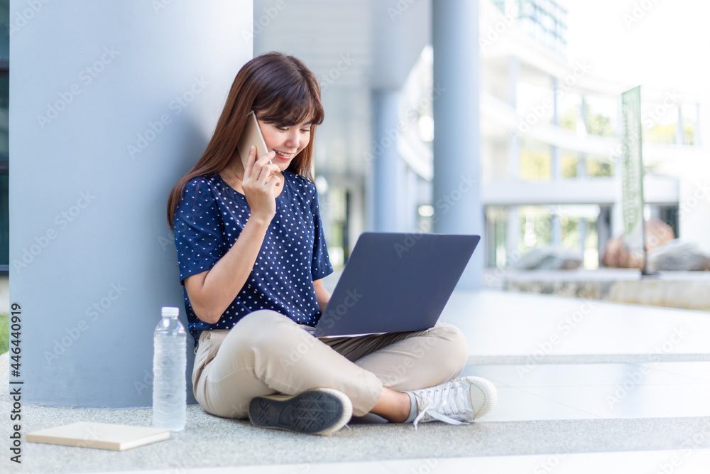 Happy young Asian woman sitting and talking on her phone while using her laptop computer outside of her office building Happy young Asian woman sitting and talking on her phone while using her laptop computer outside of her office building