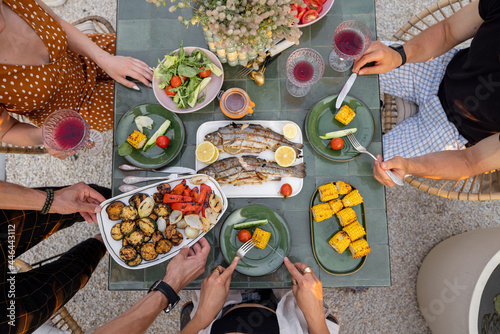 People eating at beautifully served table with grilled vegetables and fish decorateed with field flowers outdoors, top view