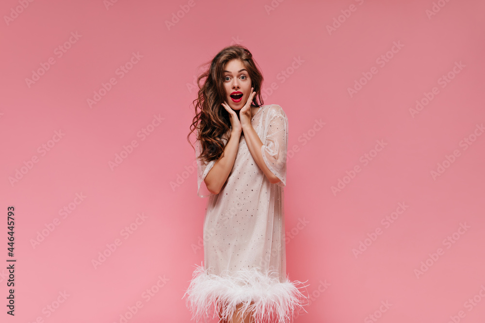 Curly young girl in white dress looks into camera in surprise and poses on pink background. Portrait of woman in stylish outfit on isolated. Curly young girl in white dress looks into camera in surprise and poses on pink background. Portrait of woman in stylish outfit on isolated.