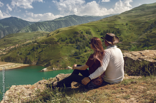 A couple sitting in front of beautiful lake and and high mountains and relaxing outdoors. Romantic hiking concept. Family wellbeing. Toned desaturated image.