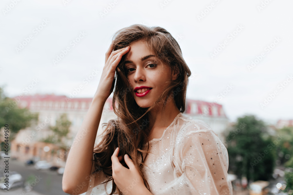 Pretty woman in white dress with sparkles looks into camera. Brunette curly girl with red lips poses on balcony outside and smiles. Pretty woman in white dress with sparkles looks into camera. Brunette curly girl with red lips poses on balcony outside and smiles.