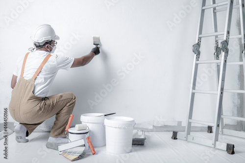 a man paints a white wall with a roller. Repair of the interior. Young male decorator painting a wall in the empty room, concept builder or painter in helmet with paint roller over the empty room