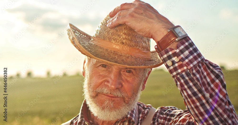 Fototapeta premium Portrait of the caucasian professional senior farmer wearing hat standing in backyard of organic farmhouse and smiling with joy. Farming business and agriculture concept