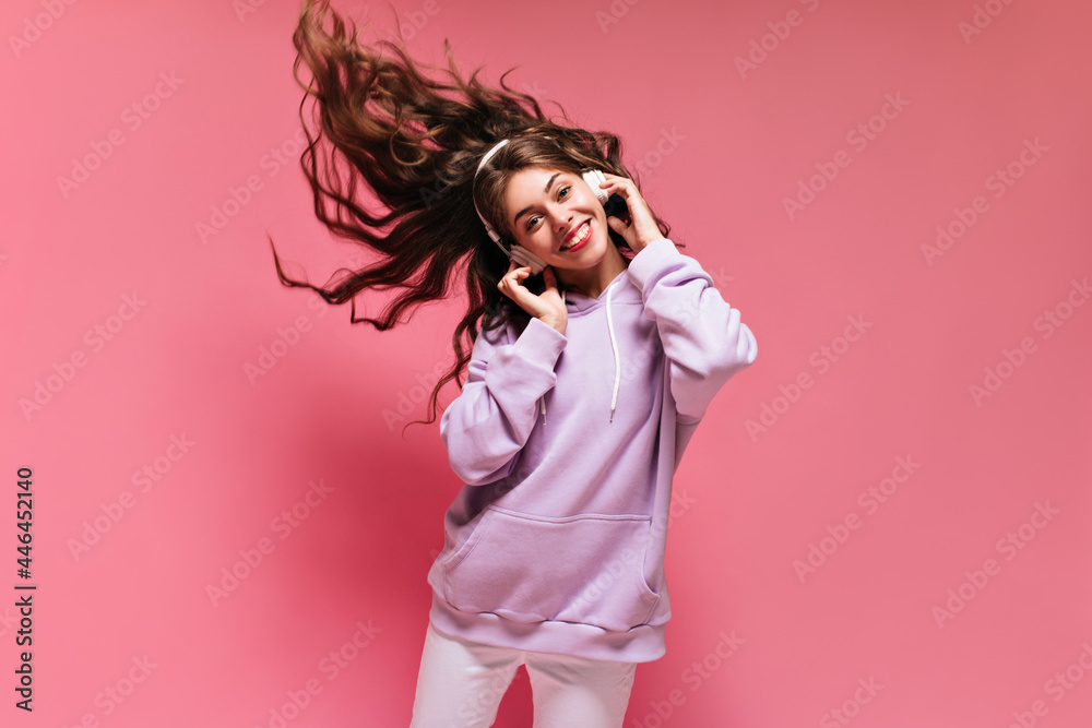 Young attractive woman plays hair on pink background. Brunette curly girl listening to music in headphones and smiling on isolated. Young attractive woman plays hair on pink background. Brunette curly girl listening to music in headphones and smiling on isolated.