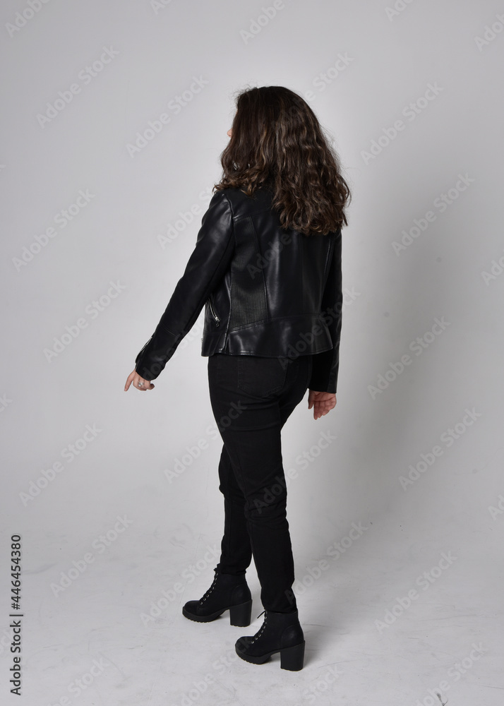 Full length portrait of young woman with natural brown hair, wearing black leather scifi outfit with jacket, standing pose with back to the camera, on light studio background. Full length portrait of young woman with natural brown hair, wearing black leather scifi outfit with jacket, standing pose with back to the camera, on light studio background.
