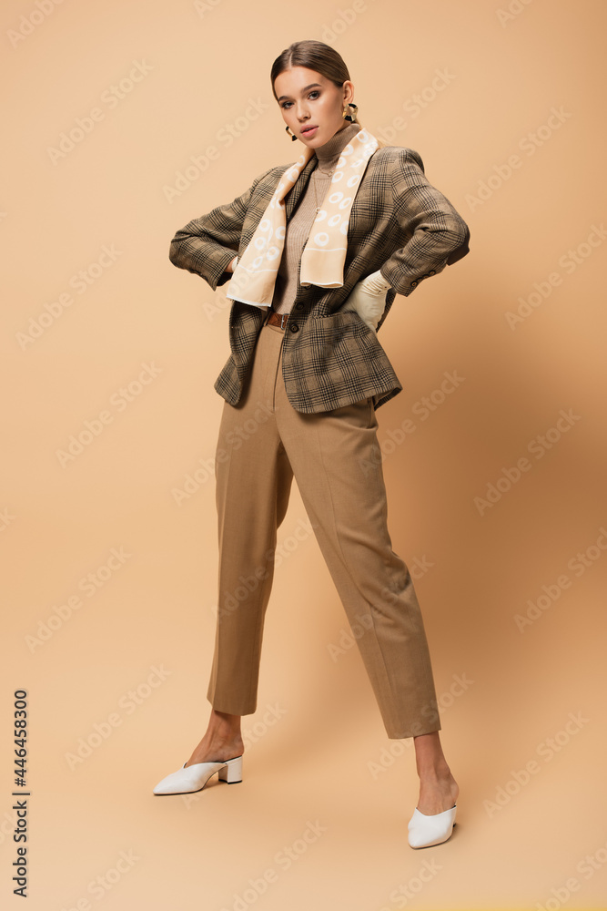 full length view of elegant woman in blazer and pants posing with hands on hips on beige full length view of elegant woman in blazer and pants posing with hands on hips on beige