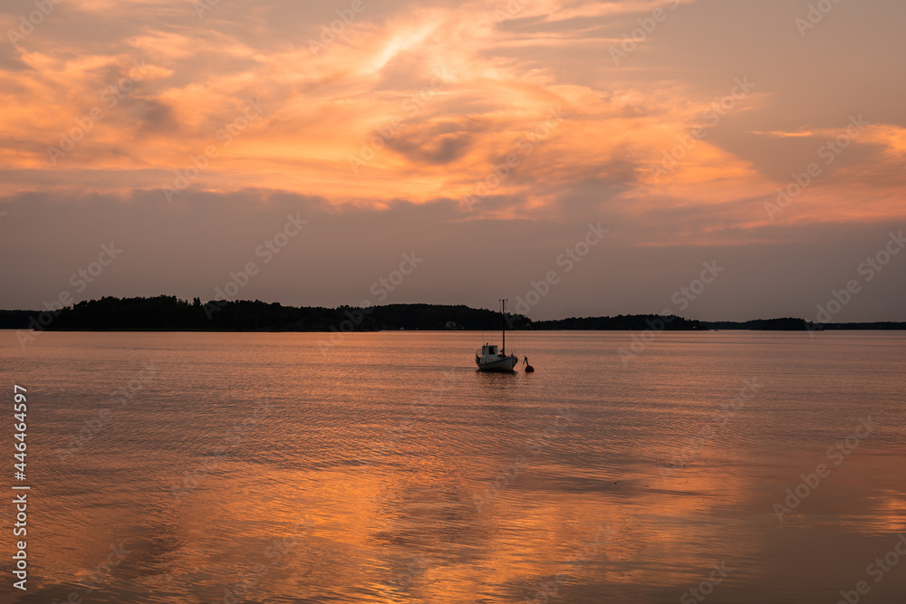 Beautiful sunset on the Baltic Sea. Amazing golden orange pink tones of the sunset cloudy sky. Islands of archipelago reflections on water surface. Small fishing boat is anchored off the coast.
