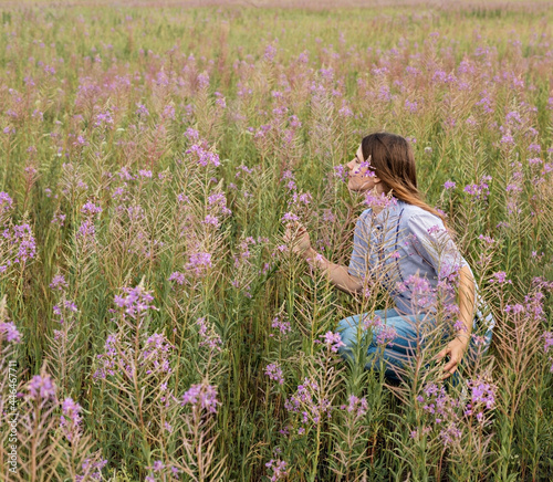 Fototapeta Naklejka Na Ścianę i Meble -  Beautiful smiling blond young woman in light purple shirt sitting on field of fireweed flowers sniffing the scent of pink flower copy space enjoing the nature