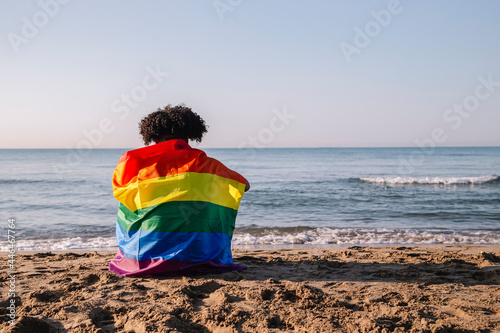Young latino man with afro hair sitting on the shore of the beach with a lgtbi flag